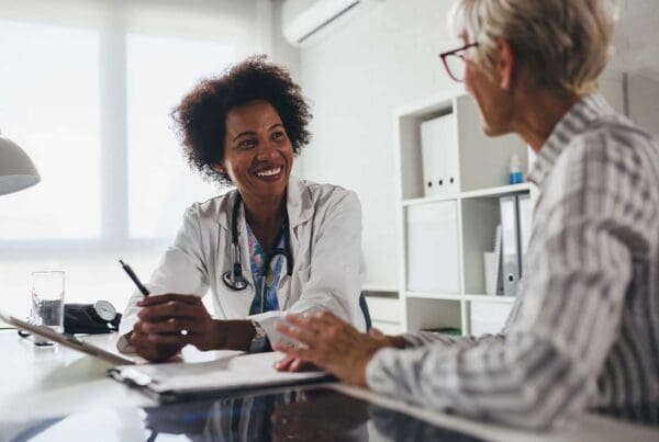 Medical Advantage Plans - Smiling Young Doctor Sits at Her Office Desk and Talks with an Elderly Female Patient about Her Healthcare Options