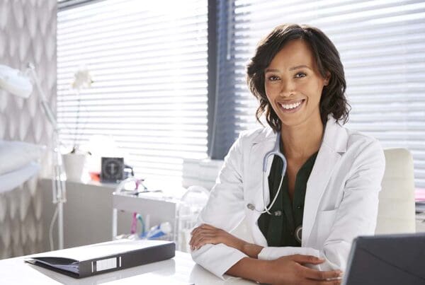 Medical Office Insurance - Portrait of Smiling Female Doctor Wearing White Coat with Stethoscope Sitting behind Her Desk in the Office